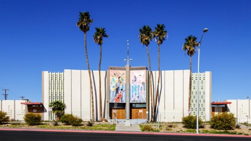 The Reed Whipple Cultural Center is a tall white building flanked on each side with green panels on the left and right in an alternating fashion. The center of the building has a mural-like pastel paintings of musicians and art on each side, split in half. The front doors are wooden and overlook the street. There are five palm trees located in front of the building.