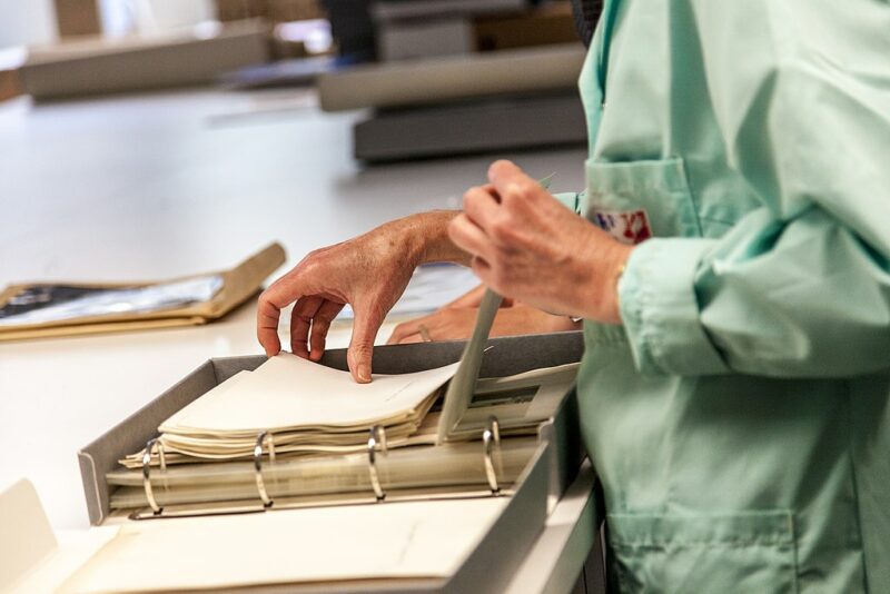Close up of the hands of an archivist going through papers in a binder in an archival box resting on a table. 