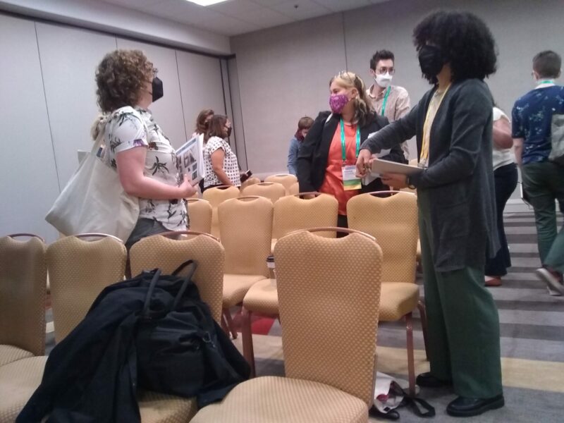 Several masked working group attendees are moving around the room to gather in small groups during the session. They are facing each other and rows of beige chairs are visible,