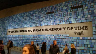 Long, decorative wall with alternating shades of blue-colored tile at the National 9/11 Memorial. The text on the wall reads in all caps, "No Day Shall Erase You From the Memory of Time. - Virgil." There are people walking around the memorial wall. 