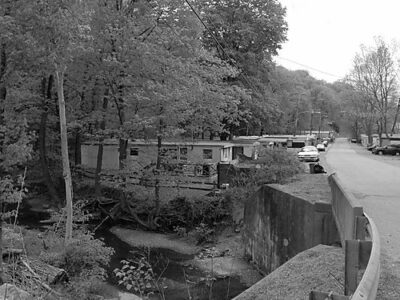 Black and white photo showing trees in the foreground, trailers in the background, and a road to the right of the image.