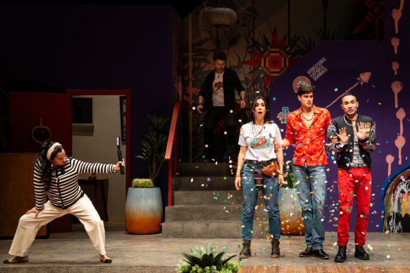 Two women and two men side-by-side in a theater set facing the audience with one man on the steps behind them. Confetti floats in the air.