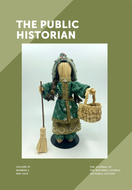 Image of corn husk doll, wearing a cap. In the right hand the doll holds a broom. In the left hand, the doll holds a woven basket by its handle.