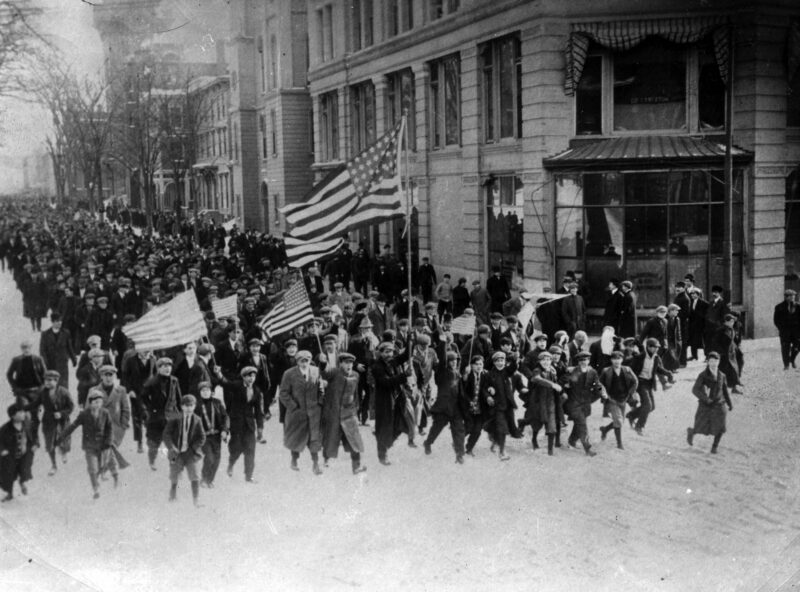 Black and white image of a large crowd marching down a main street carrying U.S. flags.