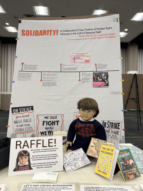 A poster board of an informational timeline from 2007 to Today that reads "Solidarity! A Collaborative 15-Year Timeline of Workers Rights Advocacy in the Cultural Resources Field!" In front of the poster board is an American Girl doll and a sign for a raffle. Three public history books sit in front of the American Girl doll.