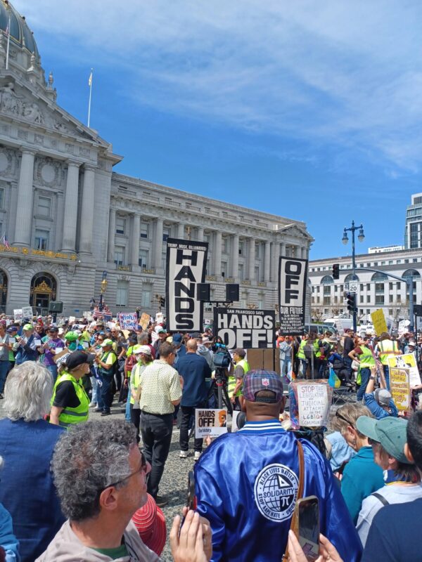 A large crowd of protesters gathers in front of a grand neoclassical government building, likely San Francisco City Hall, on a sunny day. People are holding various signs, many of which read “HANDS OFF!” in bold letters. Other signs reference opposition to attacks on the LGBTQ+ community, judicial and legislative actions, and corporate influence. One prominent sign reads “San Francisco Says HANDS OFF.” The crowd is diverse, with many people wearing yellow safety vests or colorful clothing. In the foreground, a person wears a blue satin jacket with a "Black Lives Matter – Solidarity for Justice" patch.