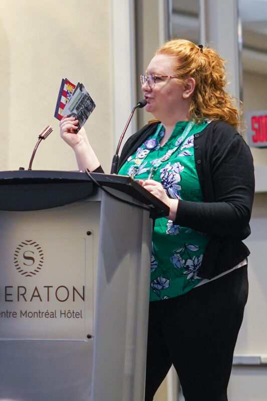 profile-view photo of the author at a podium. She is speaking and displaying a handful of NCPH "advocacy" notecards.