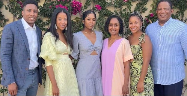 Six Black people stand in a row, their arms wrapped around each other. From left to right there is a man wearing a navy checked suit, a woman with long hair and a yellow dress, a woman with her hair up and a light blue dress, a woman with short curly hair and a pink and orange dress, a woman with her hair in braids wearing a patterned dress, and then, lastly, another man wearing a striped button-down shirt. 