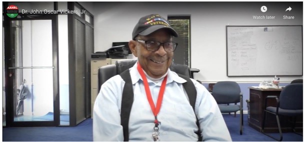 An older Black man sits smiling in a conference room. Behind him are chairs and a desk. The man wears a black cap, glasses, a red lanyard, and a button-down shirt. In the top left corner of the picture is the red and green AAHRC logo and the man's name, Dr. John Oscar Vickers Jr.