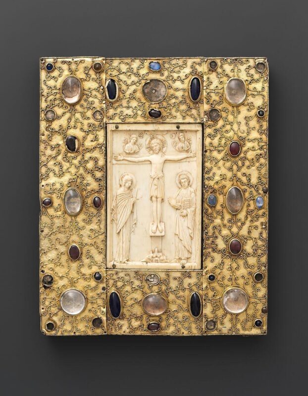 A small, rectangular gold-colored plaque coverd with filigree surrounding a rectangular ivory panel on which is carved a scene of the Crucifixtion. The cover is studded with carved glass, crystal, and numerous gemstones, including a sapphire. 