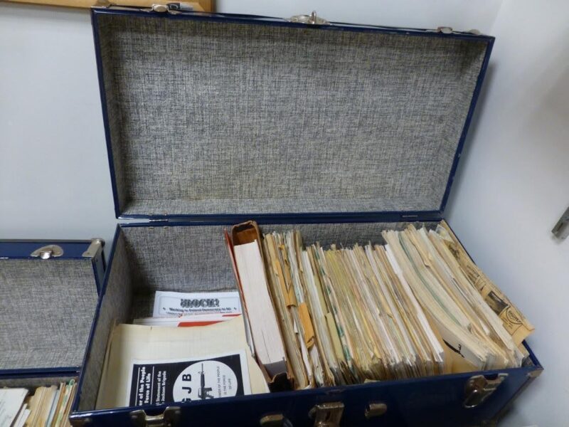 Medium blue box filled with paper material including stacks of manila folders, pamphlets, and one bound book.