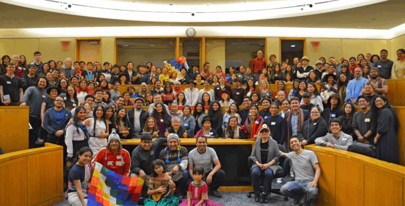 A large group of people gathered in a lecture hall posing for the camera. Several people are holding colorful textiles