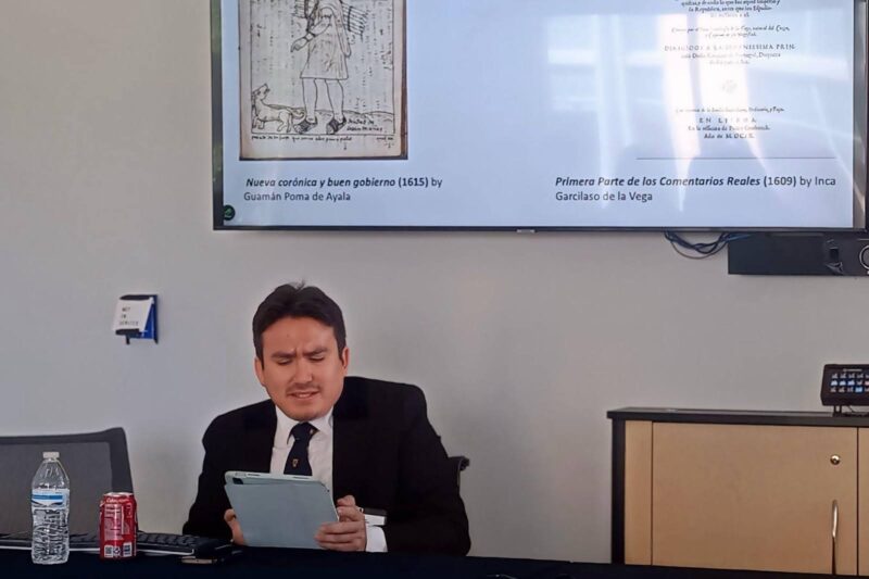 Am&eacute;rico Mendoza-Mori sits at a table in front of a slideshow projected on a screen. He reads aloud from a tablet.