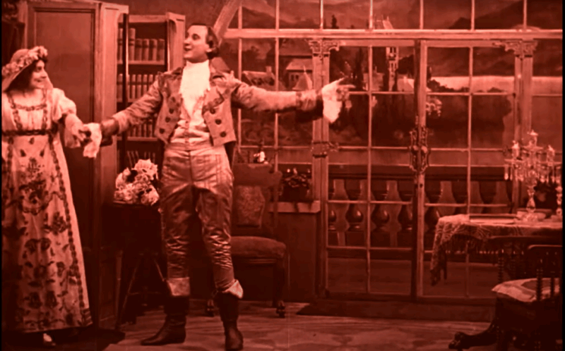 A scene of a fancy parlor with French doors. In the background are bookshelves and chairs. In the foreground a man in 18th-century clothing leads a woman by the hand. She wears a white wedding dress with floral patterns on the skirt.
