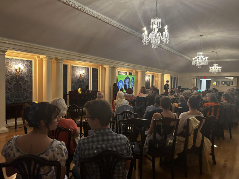 A crowd of people is seated in a large room. The walls have blue floral patterns between white columns, and the room is lit by electric chandeliers. The crowd is watching a man and woman speak in front of a TV screen showing images from a film.