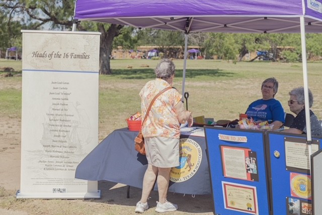 Individual standing in front of table with display on history and genealogy with two individuals sitting behind the table under a purple tent. Sign to the left of the tent reads "Heads of the 16 Families." List of family names is not legible.