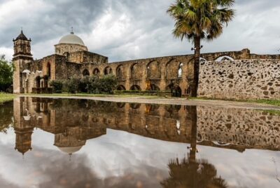 Landscape view of historic stone structure of the Mission San Jos&eacute; with water in front and a palm tree in front right