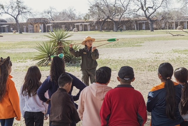 NPS ranger stands in front of a group of children doing an atlatl, a spear-thrower used for hunting by native communities in the region, demonstration as part of a school field trip. In the background is a large plant, several trees, and a line of buildings.