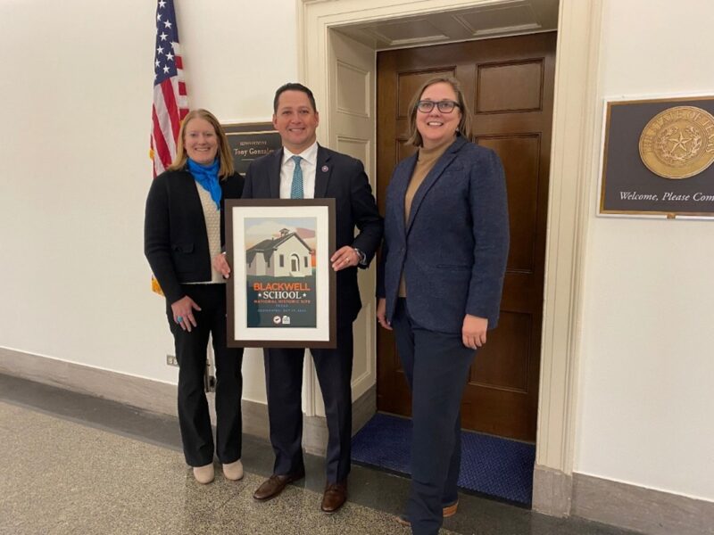 Alt text: Three people stand in a hallway, with the center person holding a framed poster and an American flag and plaques in the background.