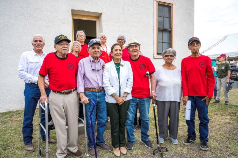 A group of older adults, many wearing red shirts, pose outside a building with a woman in the center wearing a white blazer.