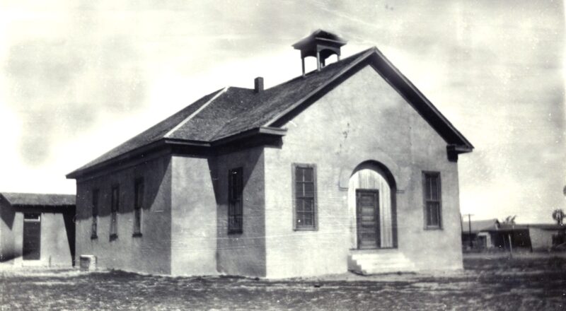 A small, old-fashioned adobe-covered schoolhouse with a bell tower and arched doorway.