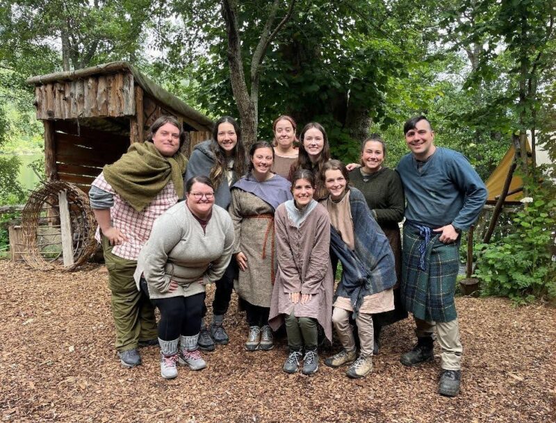 A group of ten students dressed in Iron Age costumes stand in front of trees and outbuildings.