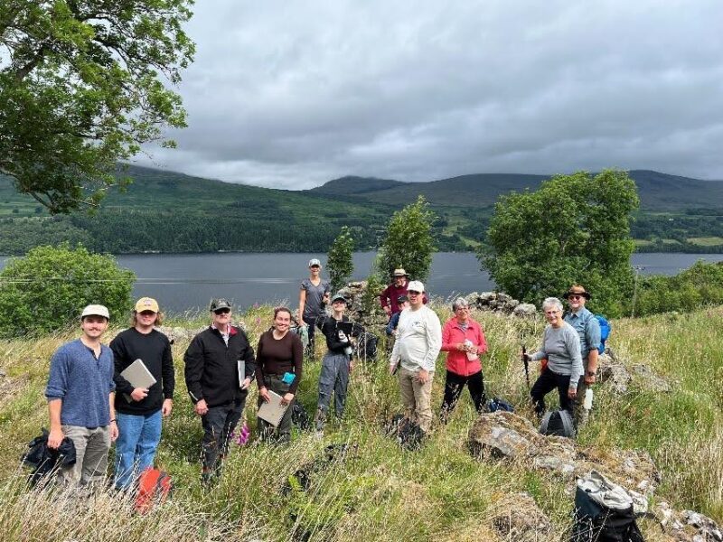 A group of eleven people engaged in fieldwork stands on a grassy hillside overlooking a lake. Green mountains appear in the background.