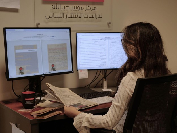 Person seated at a computer workstation with two monitors, reviewing archival materials displayed on the screens and in their hands.