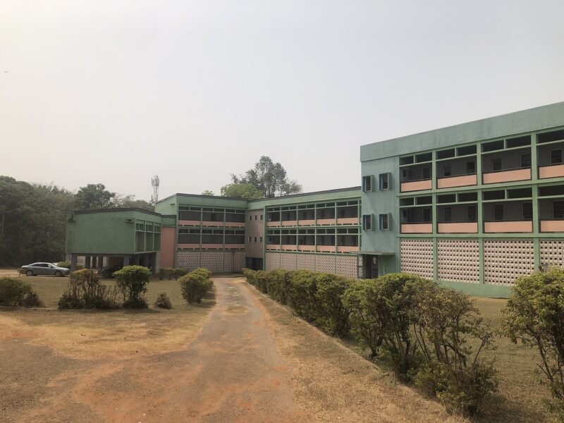 Front view of the National Archives of Nigeria building in Ibadan, a green multi-story complex with open corridors, connected wings, and a narrow driveway lined with low hedges.