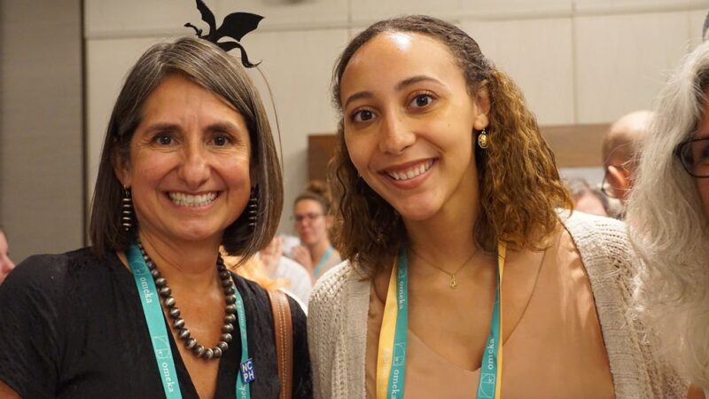 Two women smile at the camera with a reception in the background.