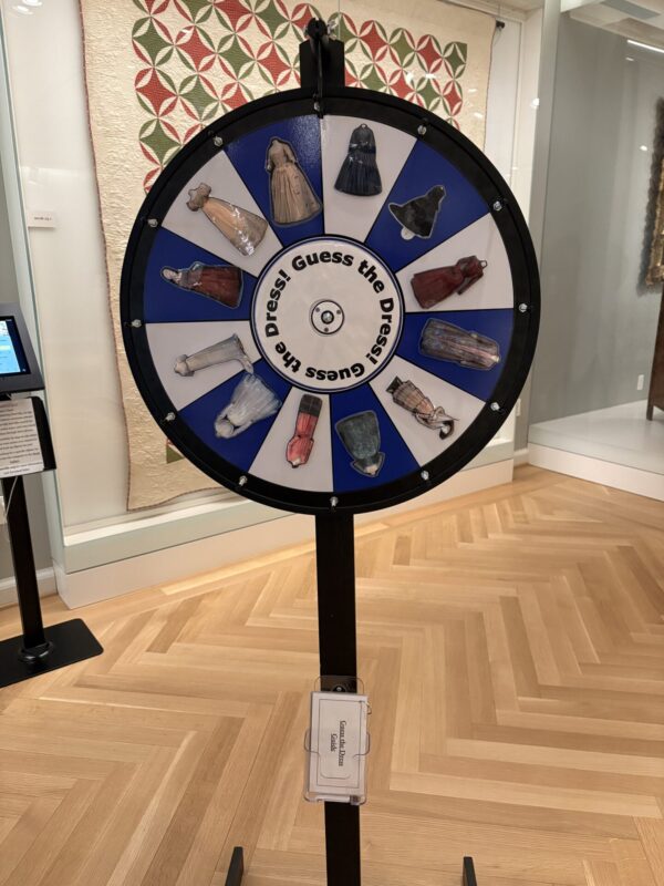 A stand holding a spinning wheel stands in front of a white quilt covered in green and red triangles. The wheel has alternating blue and white slices. On each section, there is a picture of an antique dress. Text on a white inner wheel reads: “Guess the Dress!”
